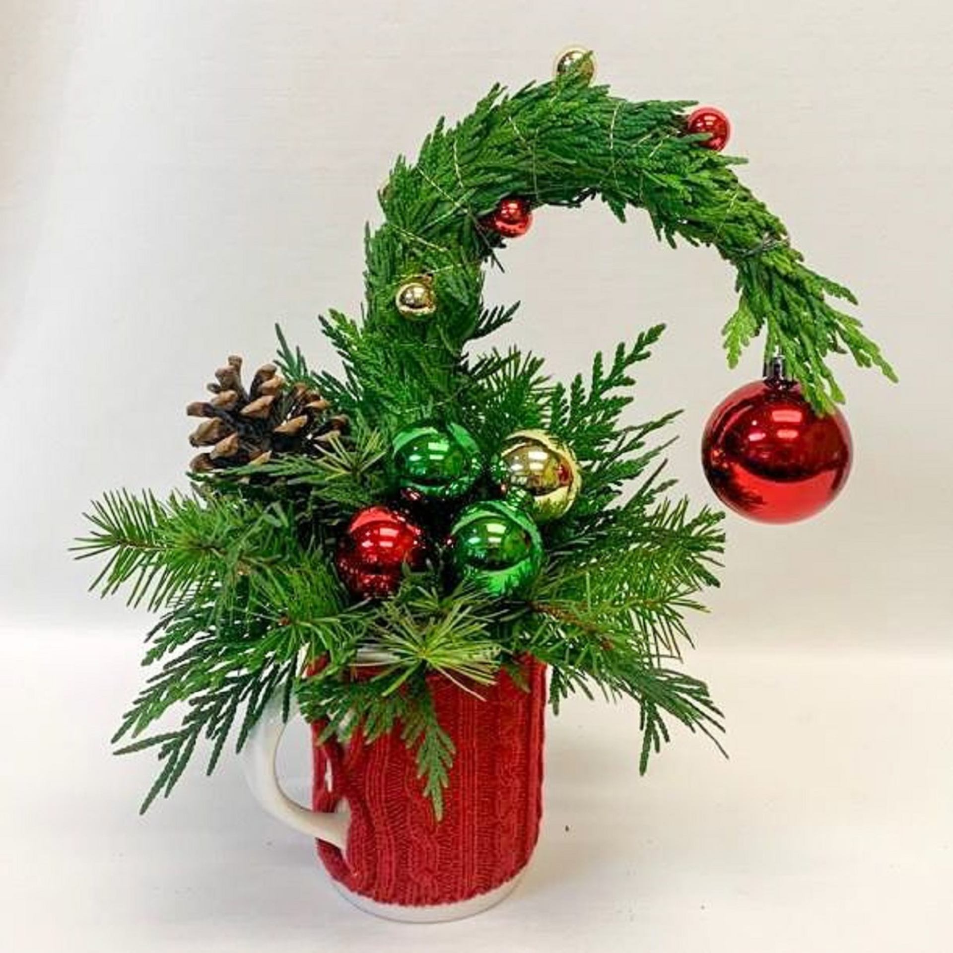Festive pine arrangement in red mug with ornaments and curved top.