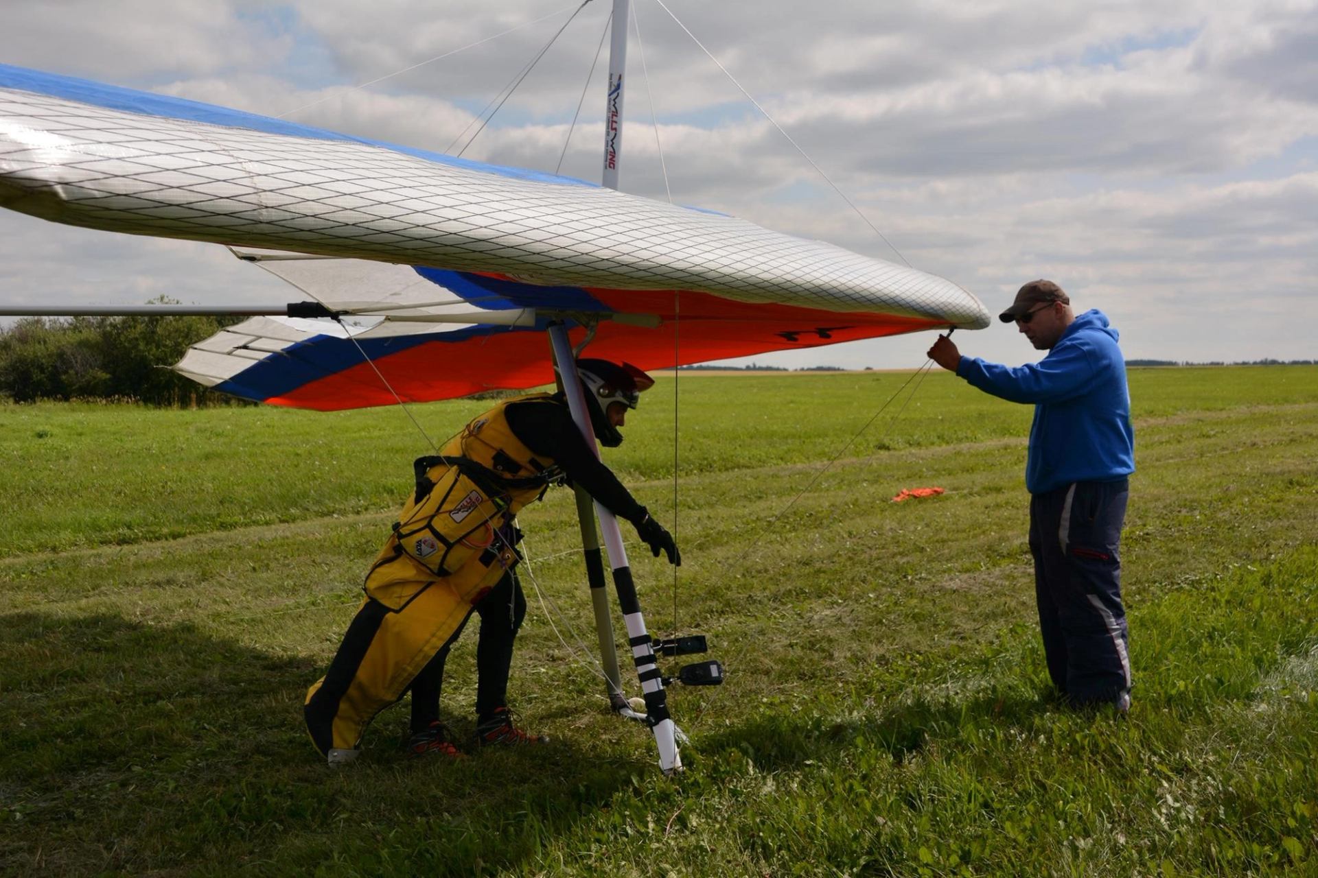 Hang glider on a grassy field with one person preparing to launch and another assisting.