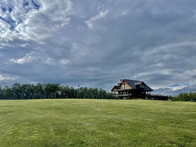Log cabin on open grassy field beneath cloudy sky and tree line.