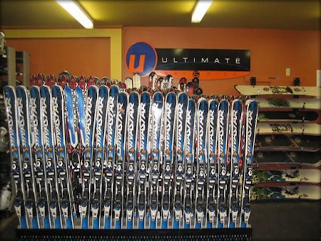 A display of colorful skis is lined up vertically in a sporting goods store, with a sign reading "Ultimate" on an orange wall in the background.
