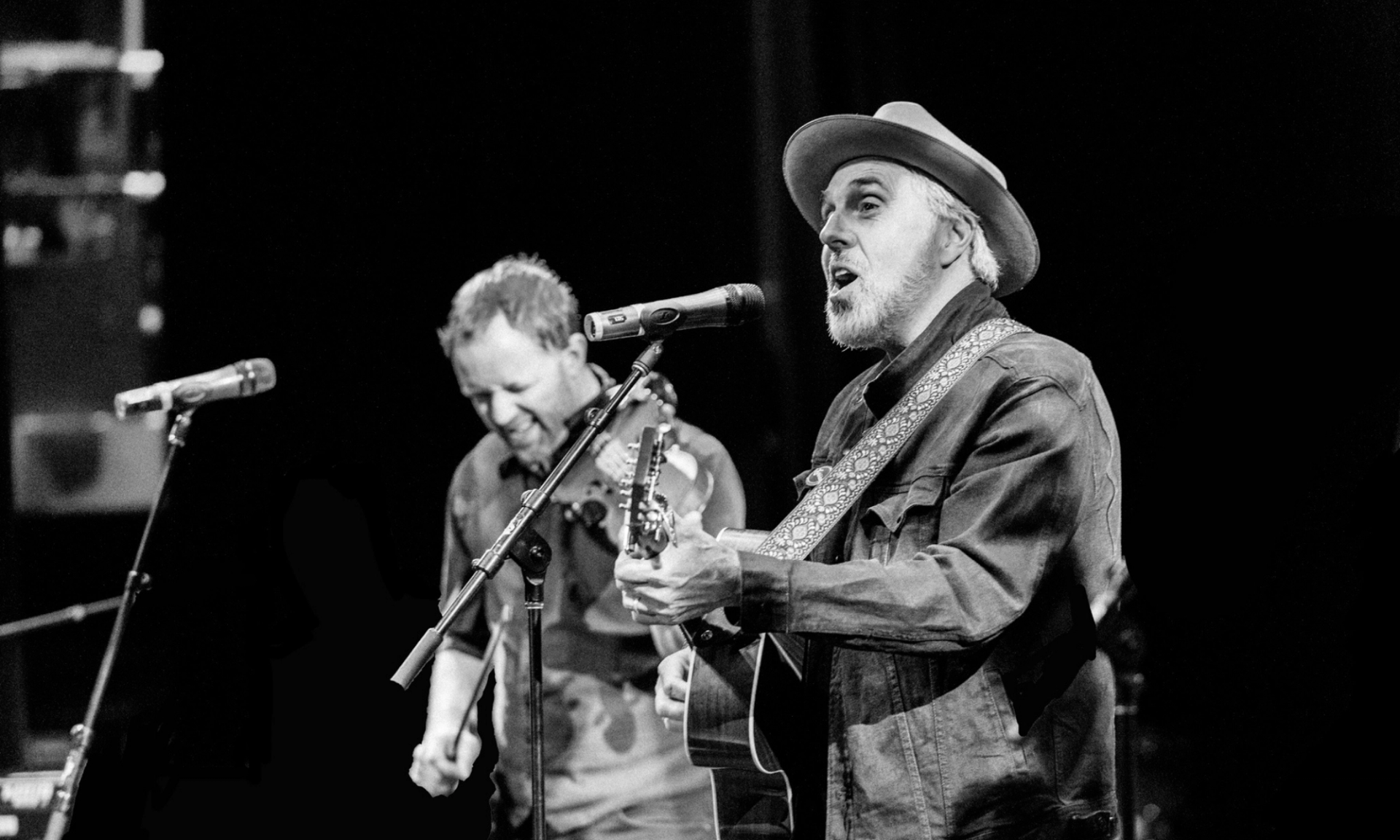 Black and white photo of two musicians performing with guitar, fiddle and microphones onstage.