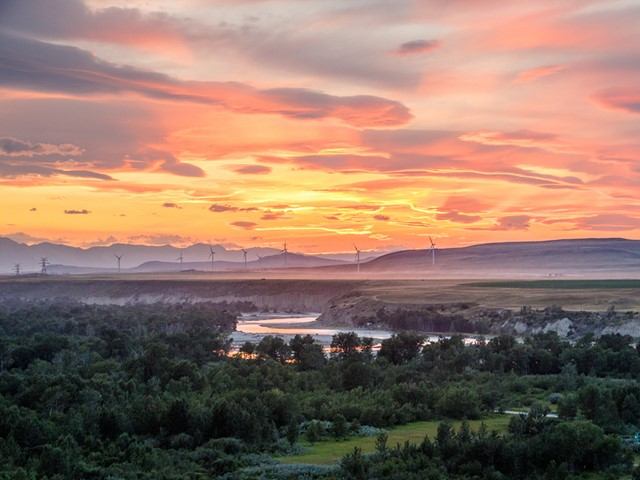 Sunset over Waterton Valley with river, wind turbines, and distant mountains under cloudy sky.