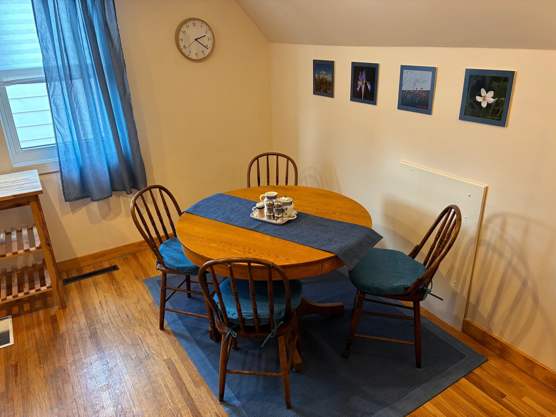 Dining nook with round wooden table, four chairs, blue accents and wall art.