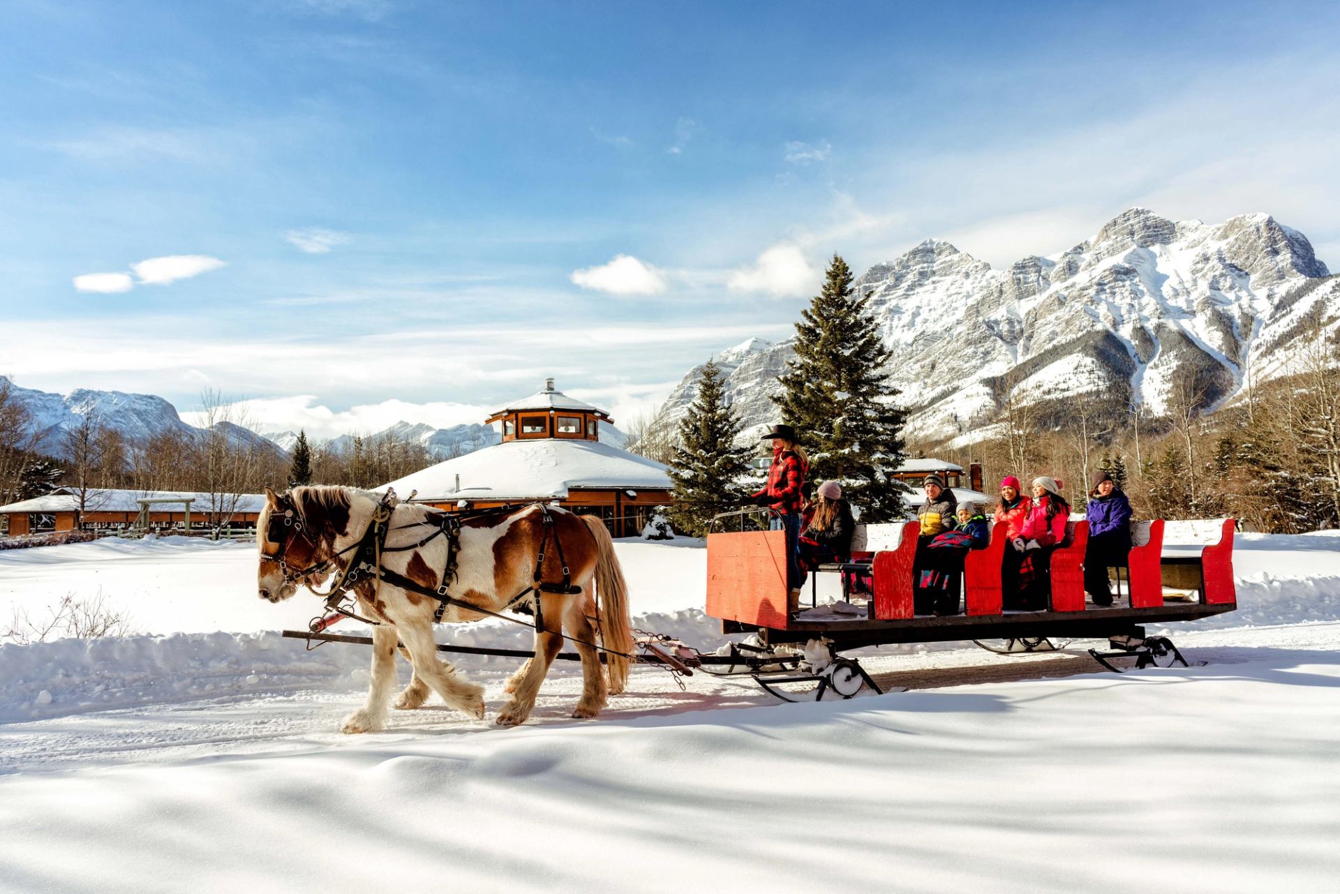 Horse-drawn sleigh carrying riders across a snowy field with Rocky Mountains in the background.