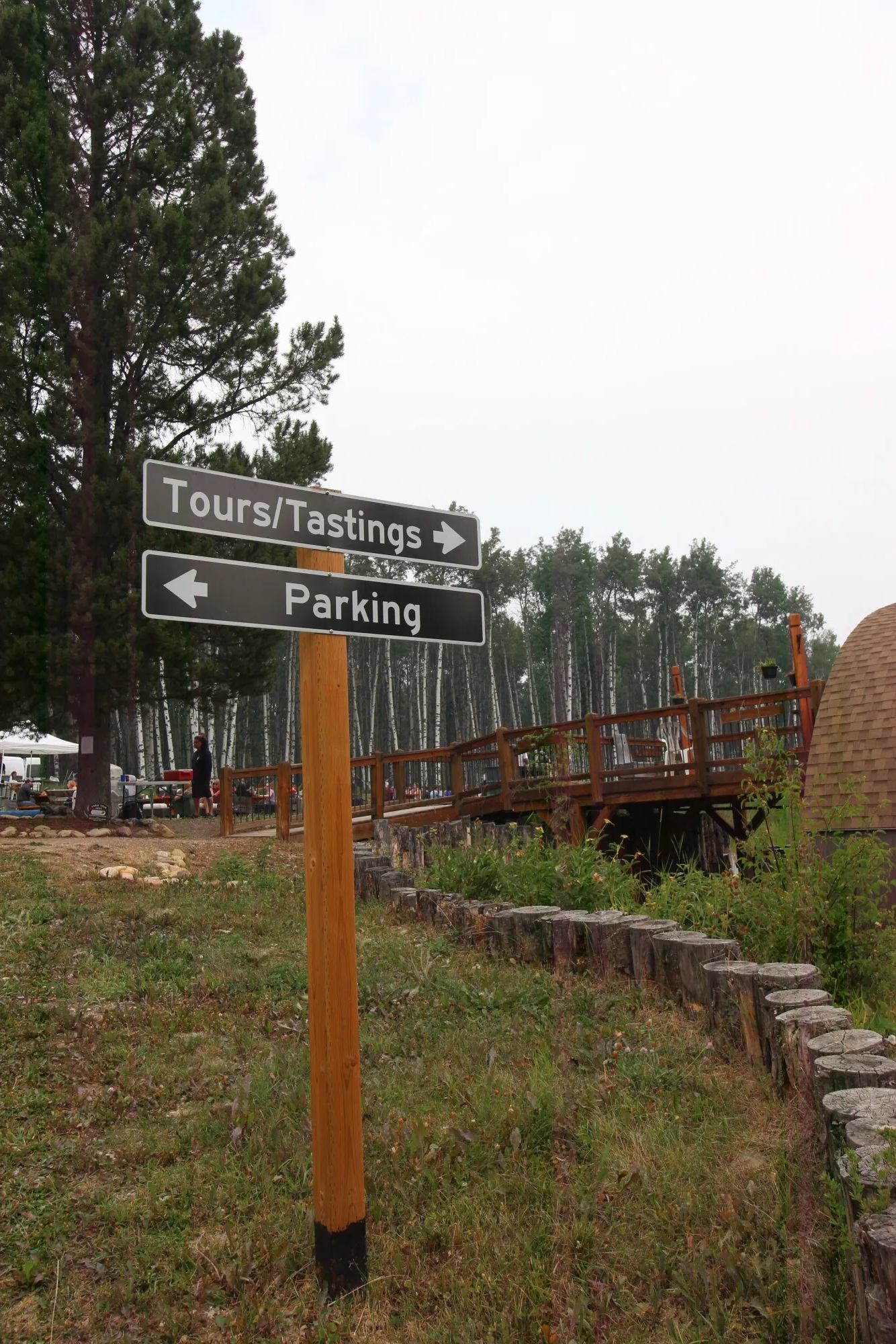 Wooden post with Tours, Tastings and Parking signs beside forested outdoor deck.