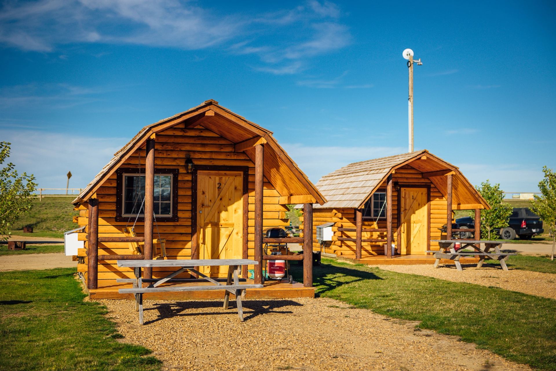 Two wooden cabins with porches, picnic tables, and trees in a grassy campground setting.