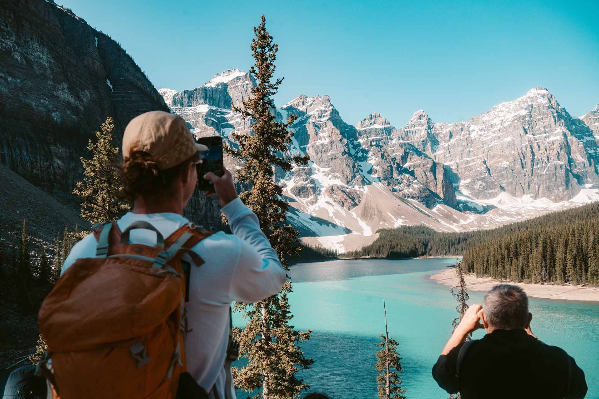 Hikers photograph turquoise mountain lake surrounded by evergreen forest and snow-capped peaks.