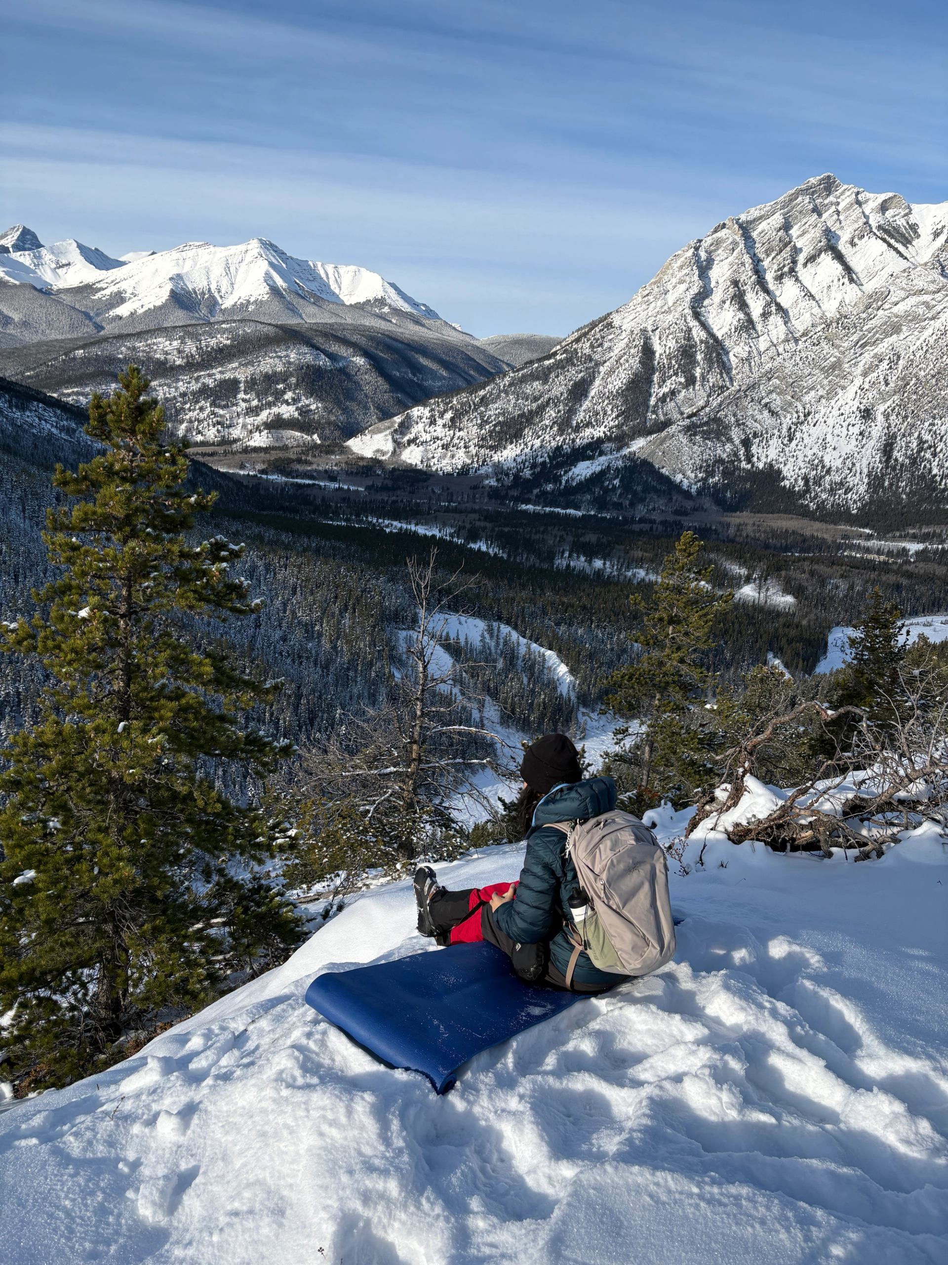 Hiker sitting on snowy slope overlooking a valley and rugged mountain peaks.