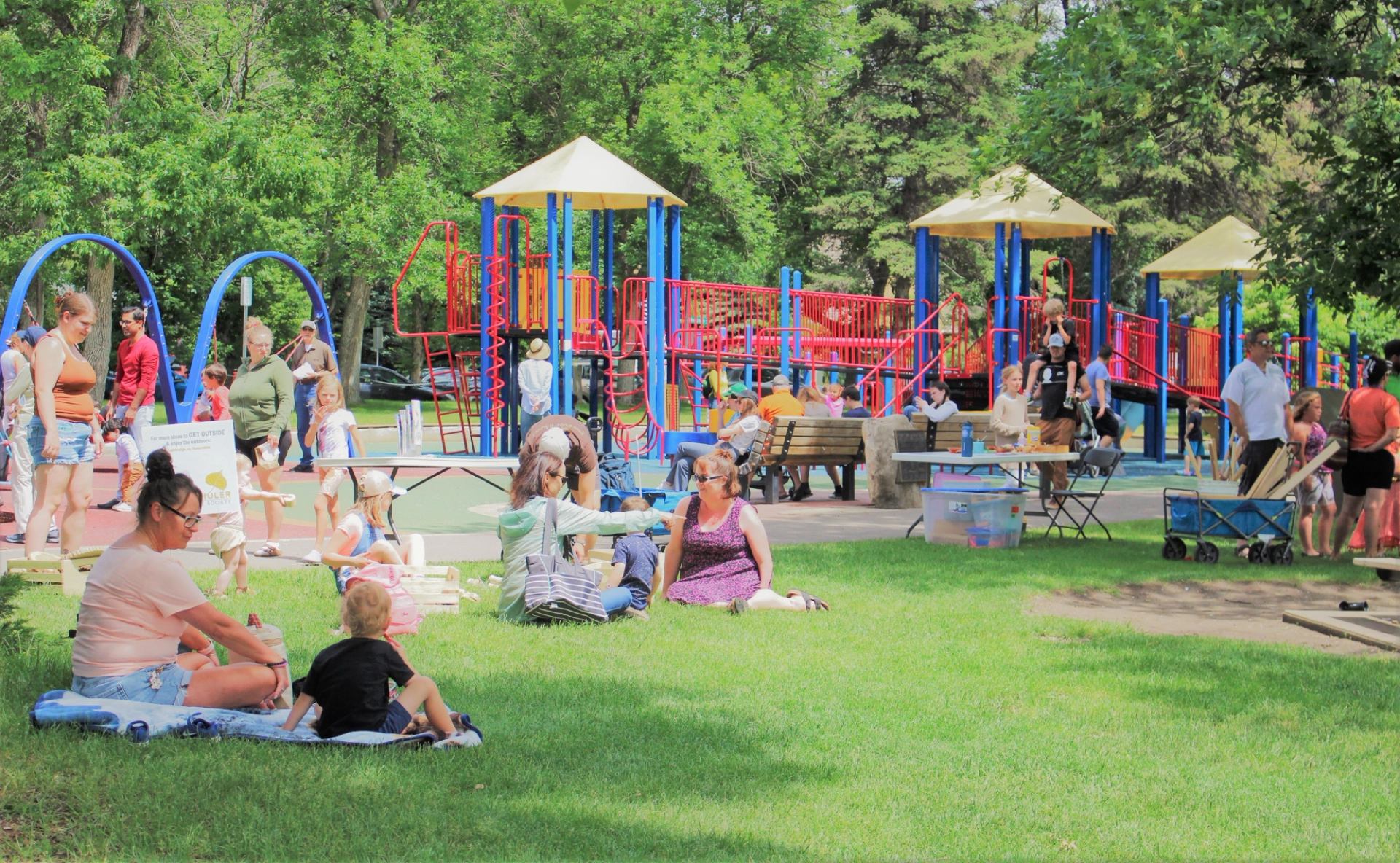 Families relax on grass while children play on colourful playground equipment in park.