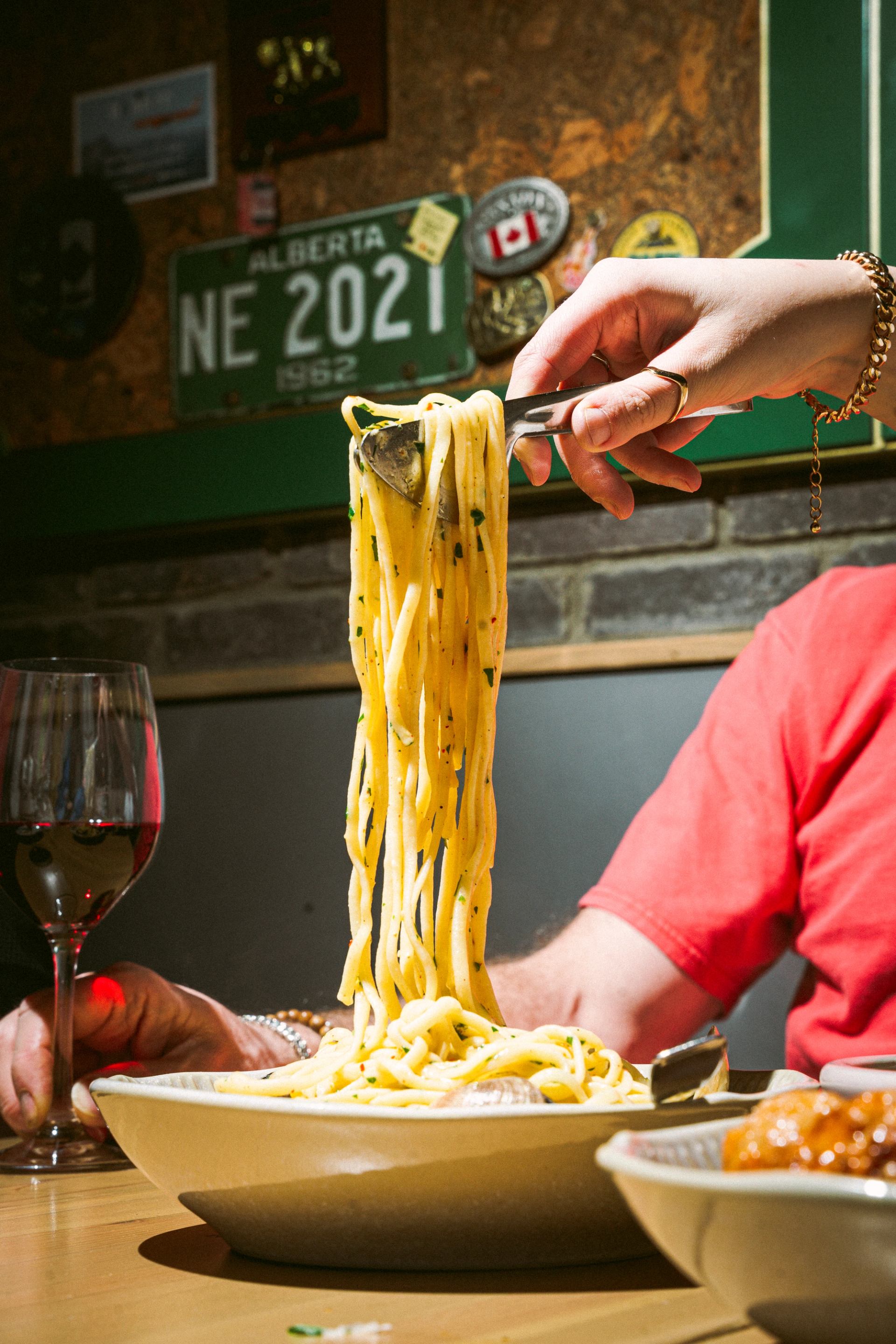A hand uses tongs to lift a large serving of spaghetti from a bowl on a table, with a glass of red wine nearby.