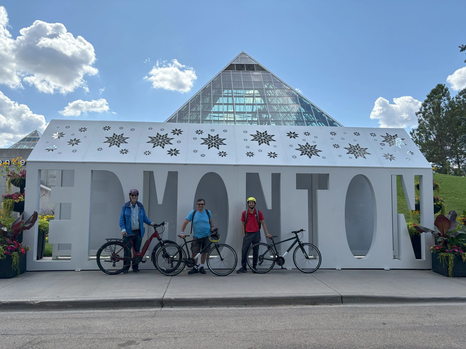 Three cyclists pose with bikes in front of “EDMONTON” sign and glass pyramid building.