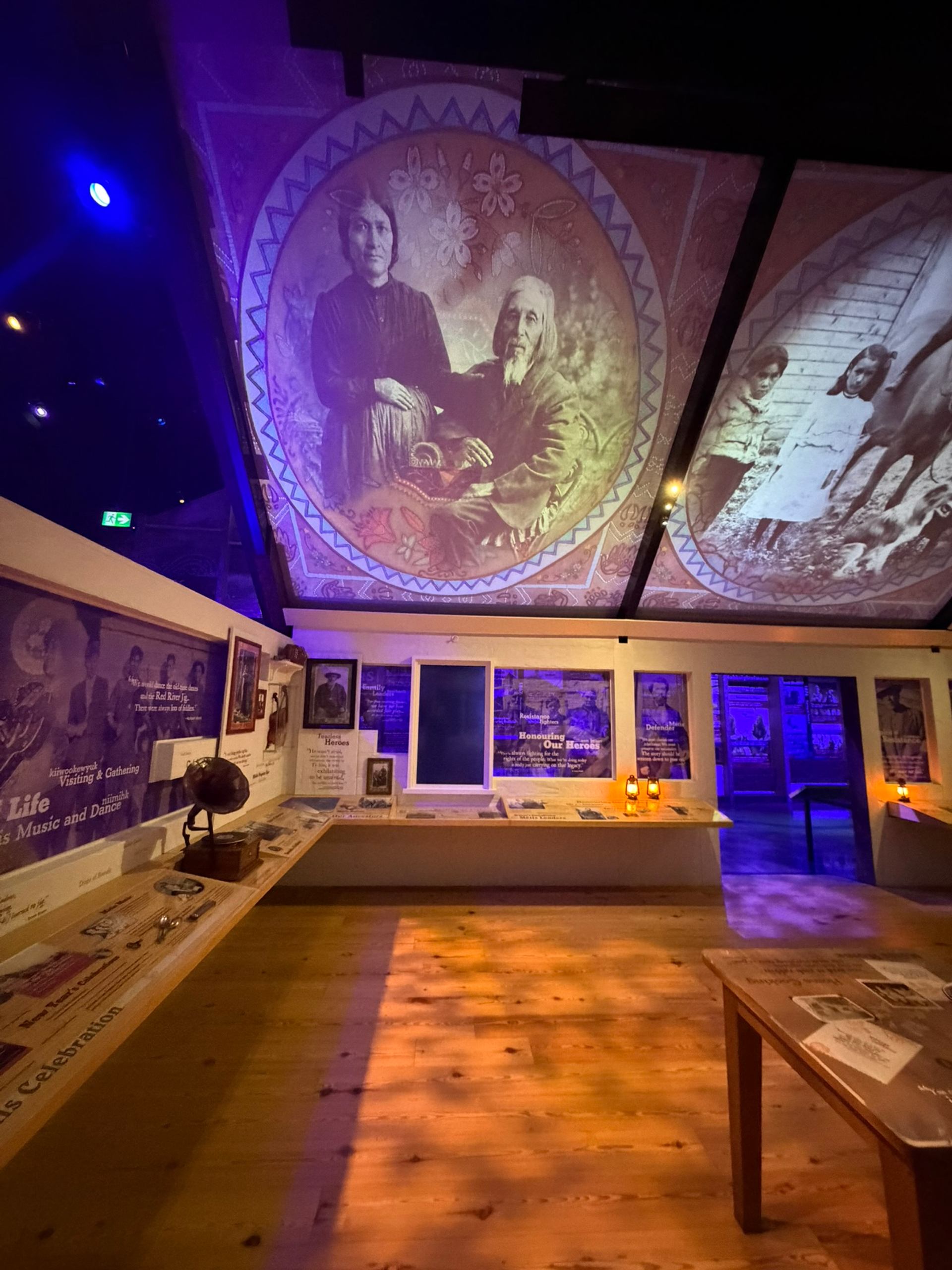 Museum exhibit room with historical photos on ceiling and display tables under purple lighting.