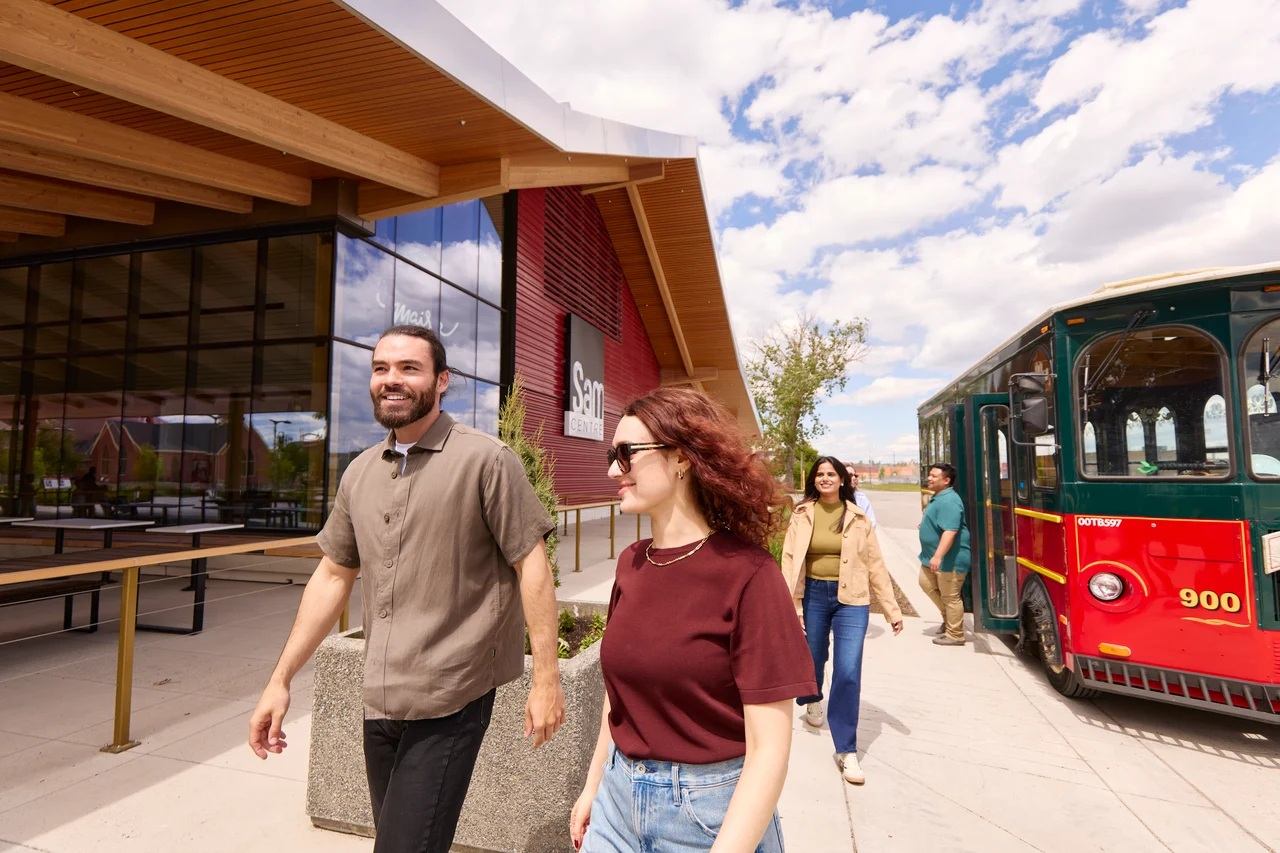 People walking on sidewalk near Sam Centre building beside red and green trolley bus.