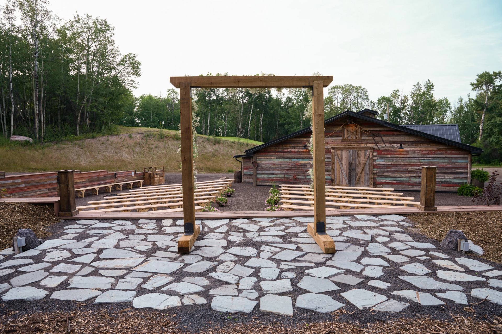 Outdoor venue with wooden benches, archway, and rustic barn in natural setting.