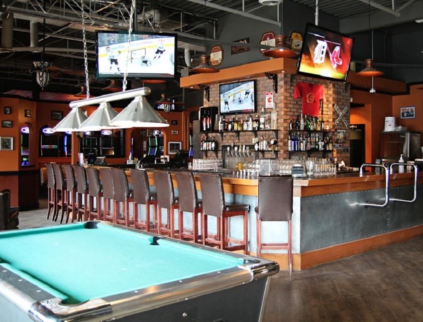 Bar interior with pool table, high chairs, TVs, and shelves of liquor bottles.
