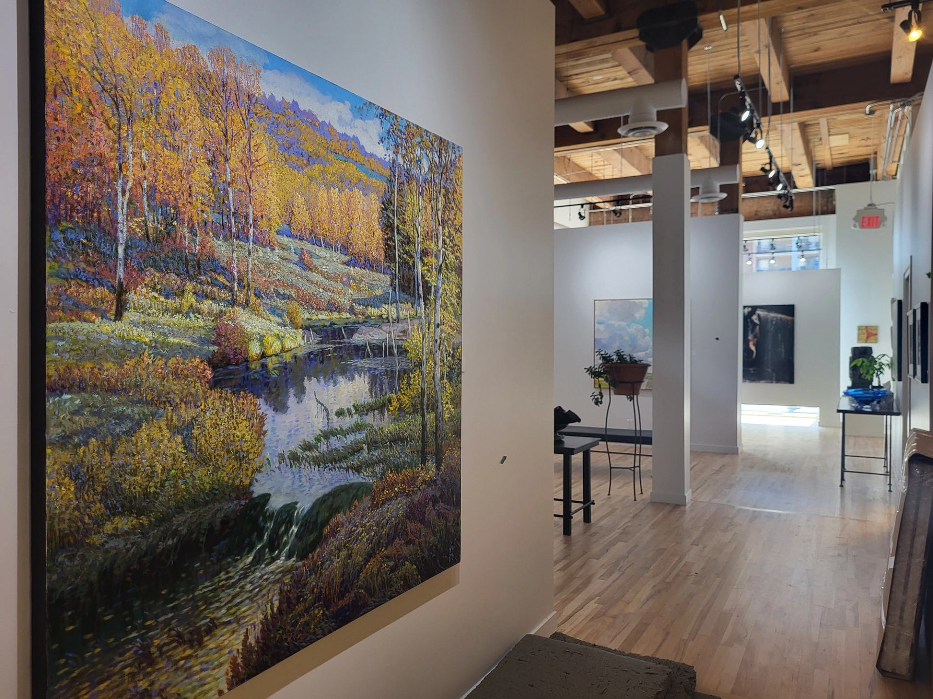 Large colourful forest landscape painting hangs in a gallery with wood floors and exposed beams.