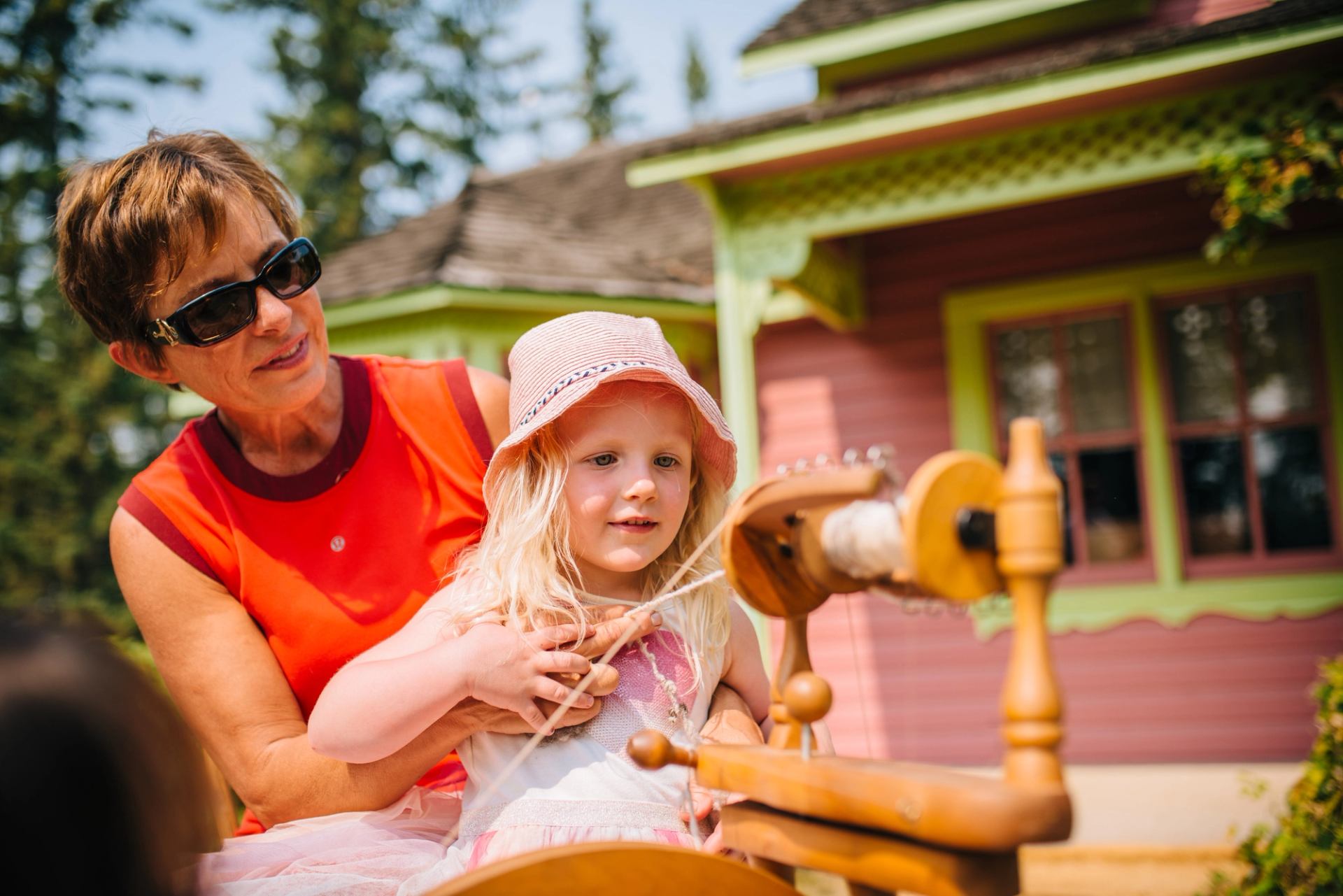 A young girl sat in a lady's arms, spinning wool, wearing a sun hat.