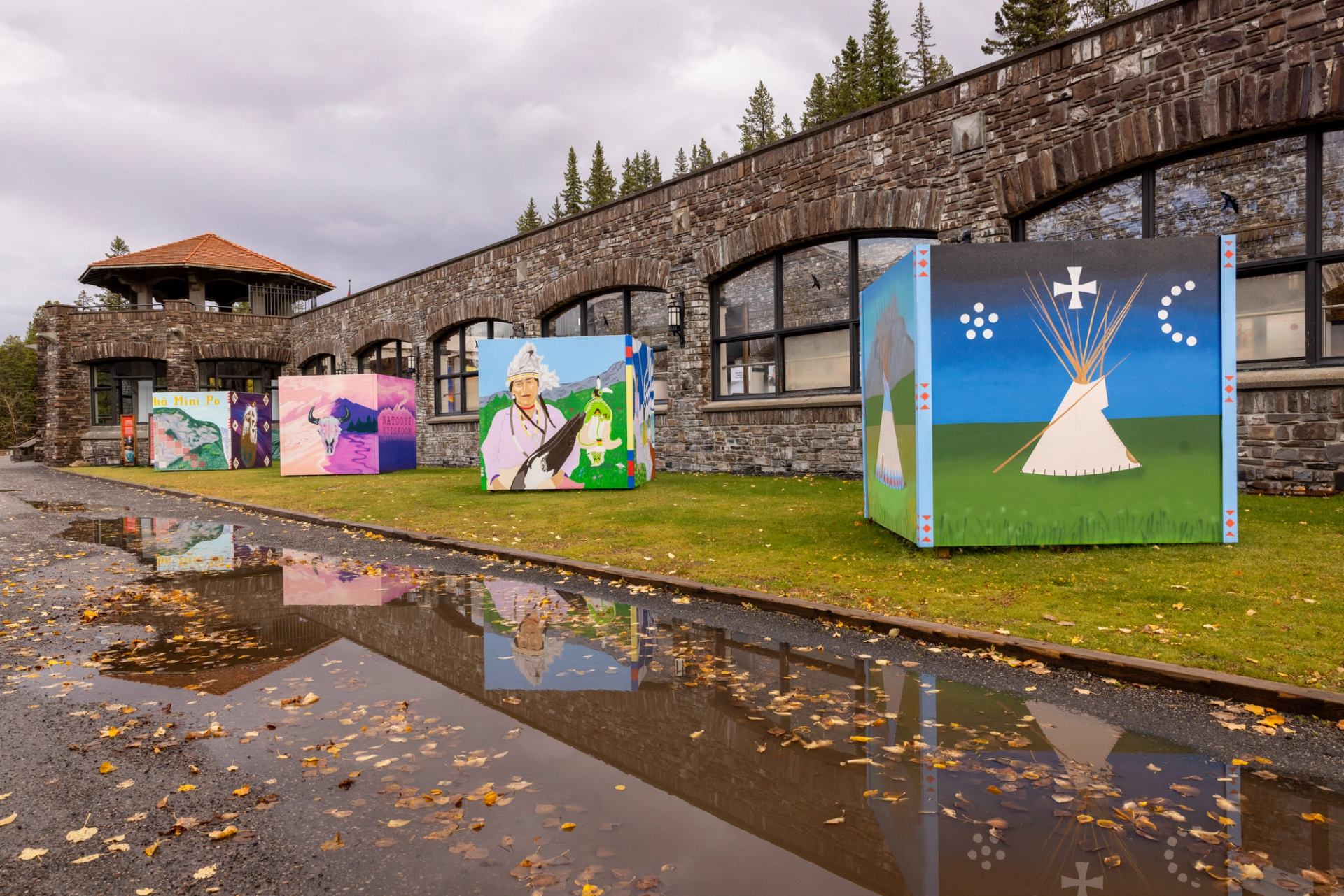 Colourful painted cubes with Indigenous-themed art displayed on grass beside a stone building.
