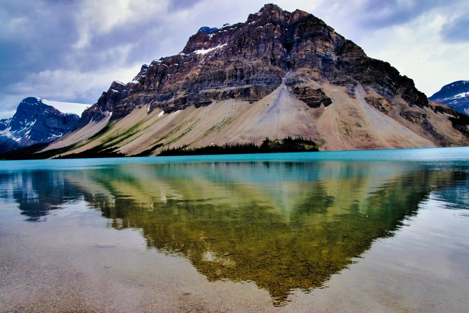 Rocky mountain with snow patches reflected in a calm turquoise lake under clouds.