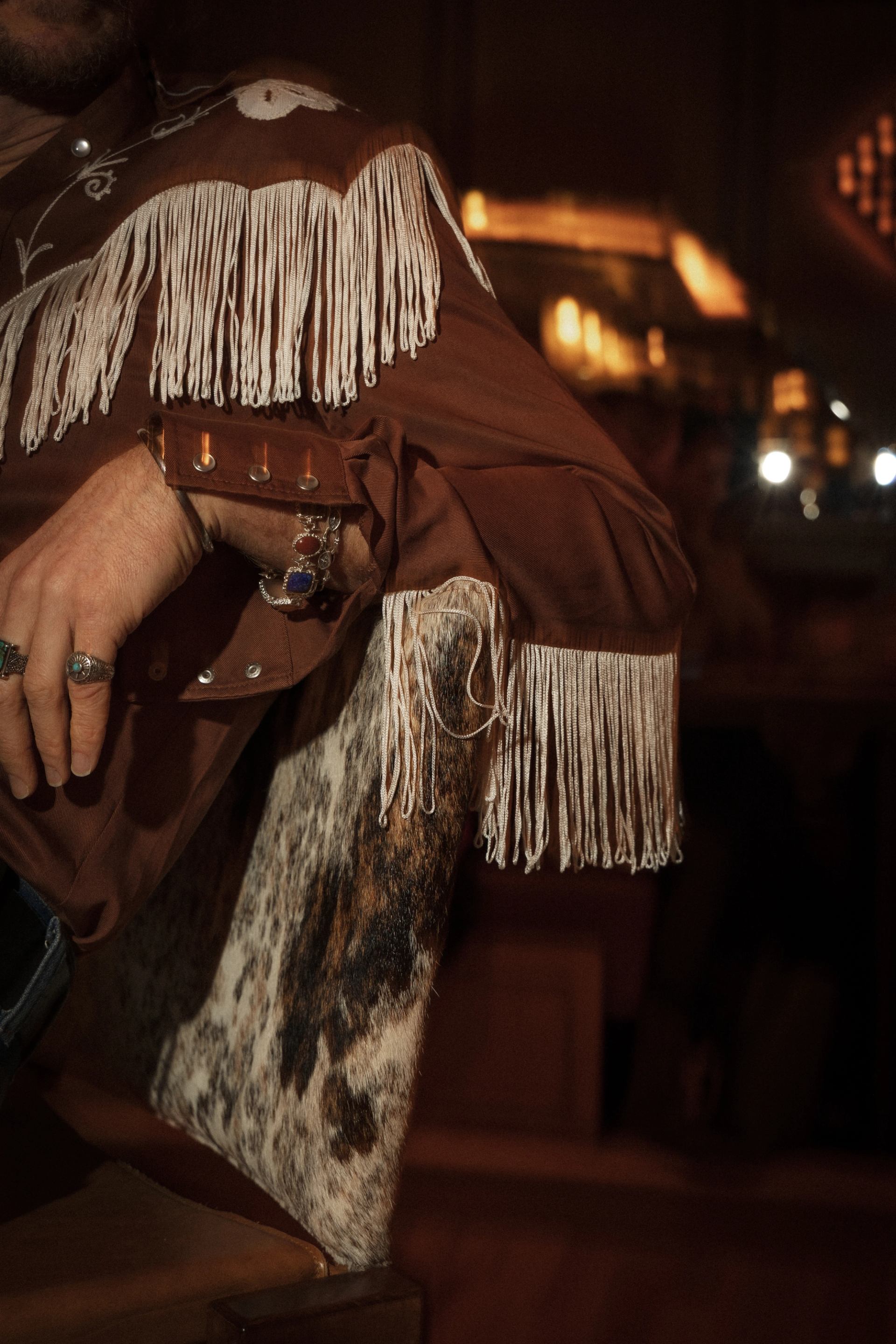Close-up of a person in a fringed western shirt resting an arm on a cowhide chair.