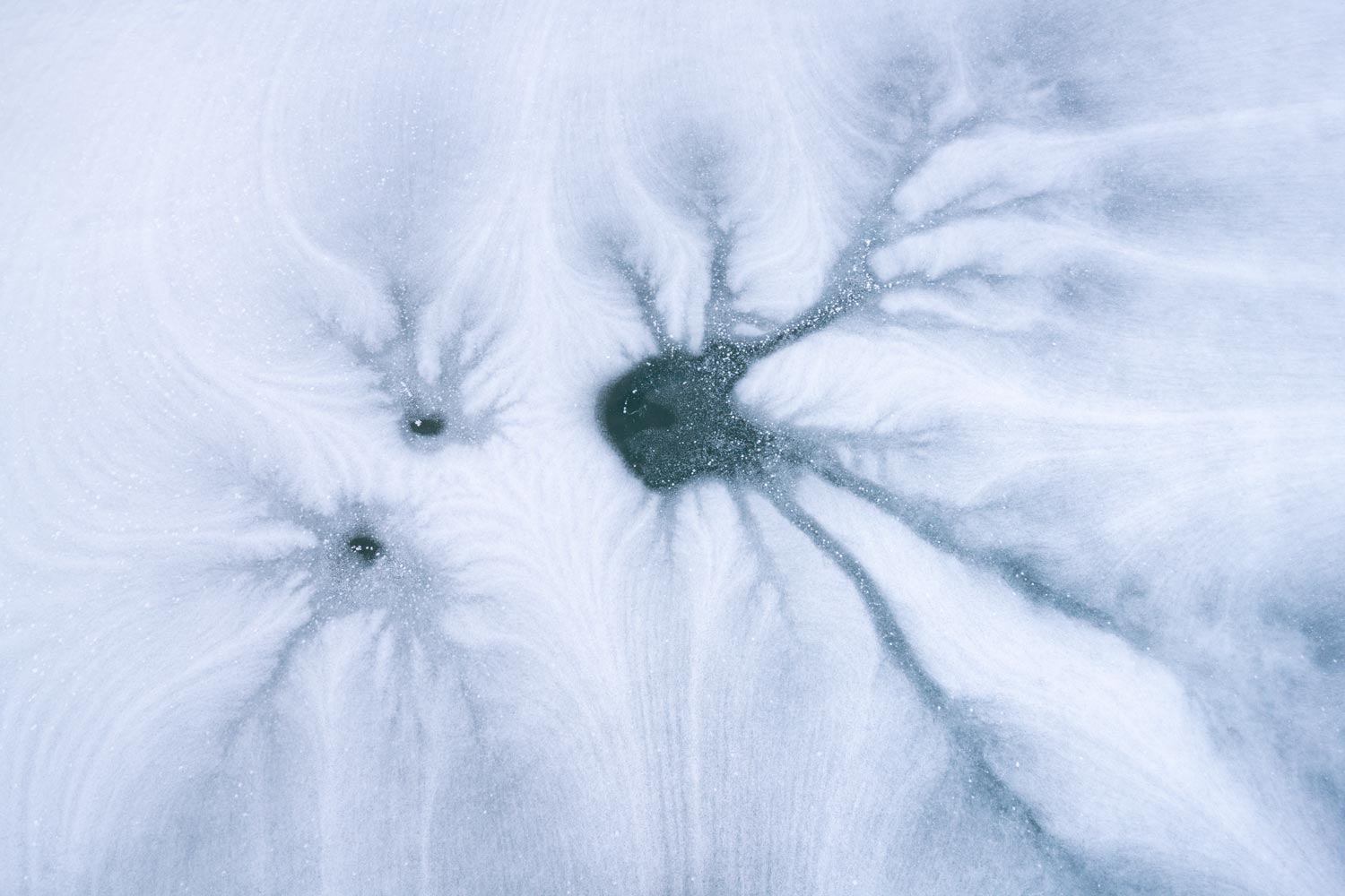 Intricate frost patterns radiating from three dark holes on an icy blue surface.
