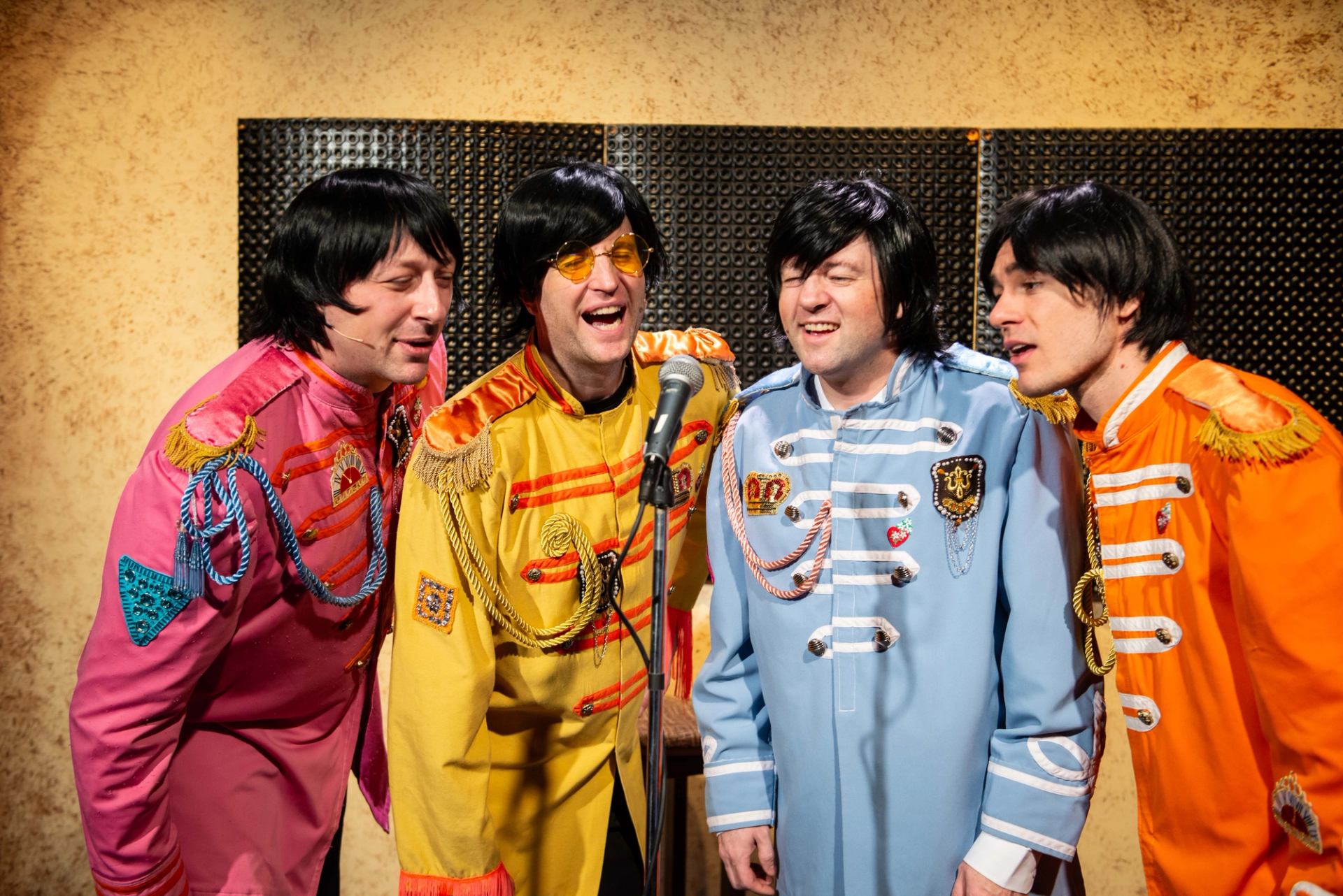 Four performers in colourful Sgt. Pepper-style uniforms sing together around a microphone.