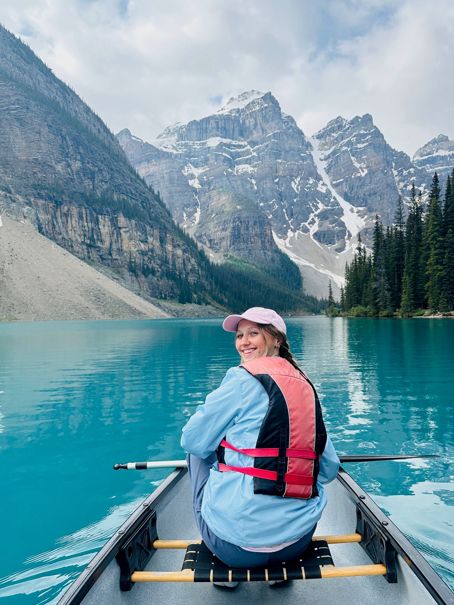 Tour boat passenger in pink cap and life jacket on turquoise lake, snowy mountains in background.