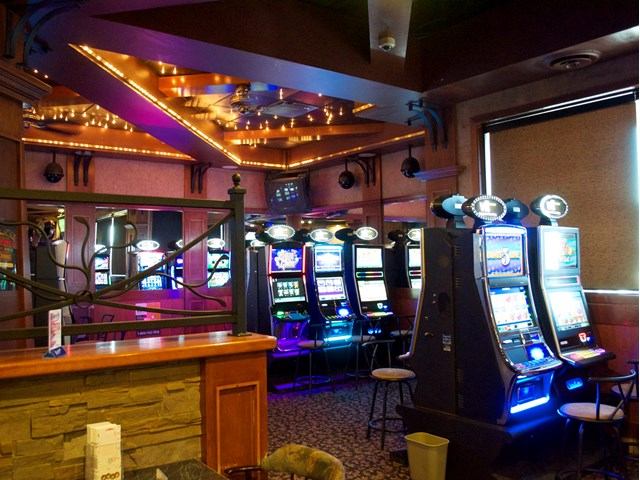 Lounge interior with slot machines, bar counter, stools and string lights on ceiling.