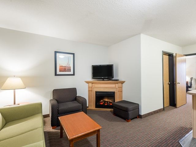 Hotel sitting area with sofa, armchair, fireplace, TV, ottoman and open doorway.