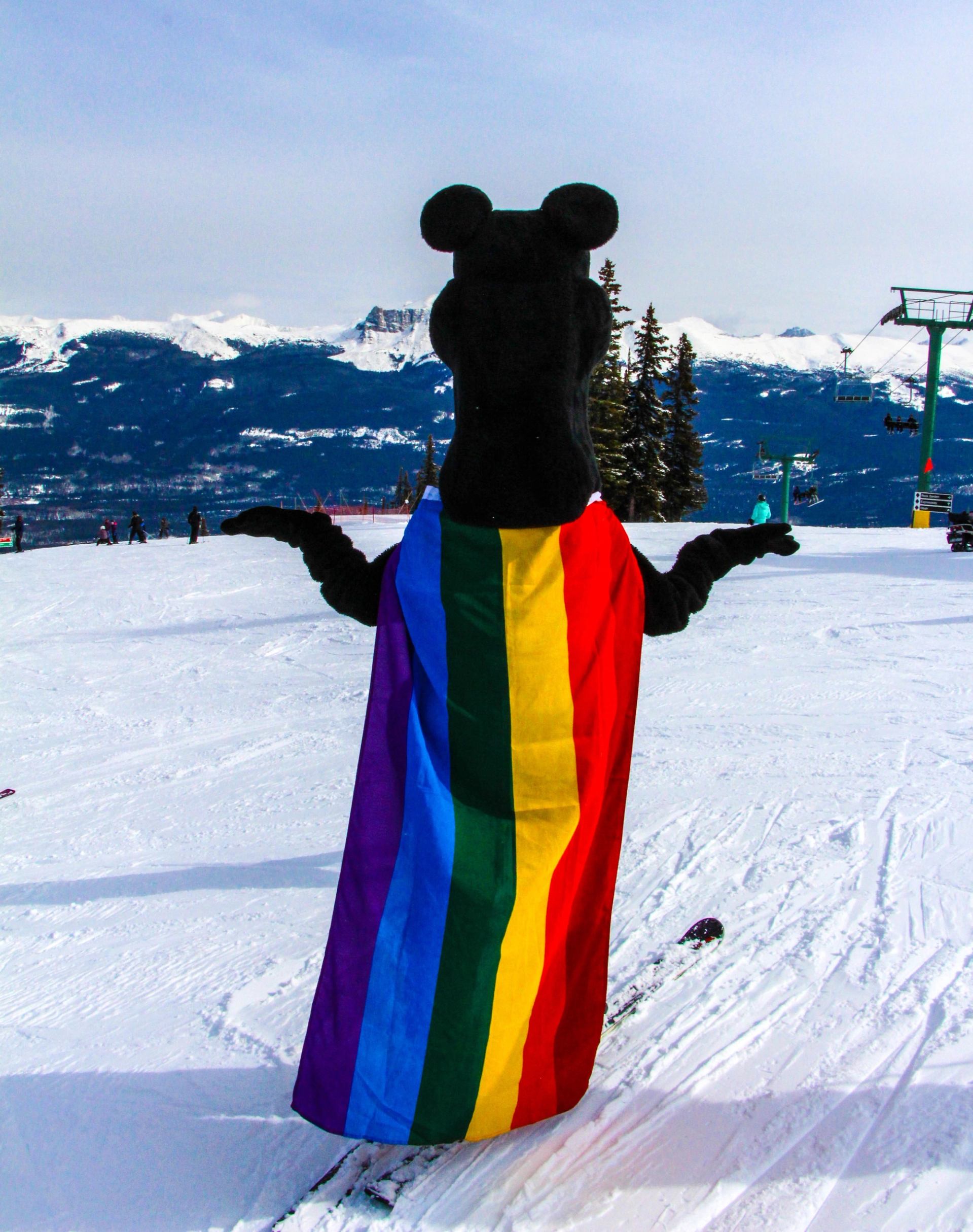 A costumed figure wearing a rainbow flag standing on a snowy ski hill.