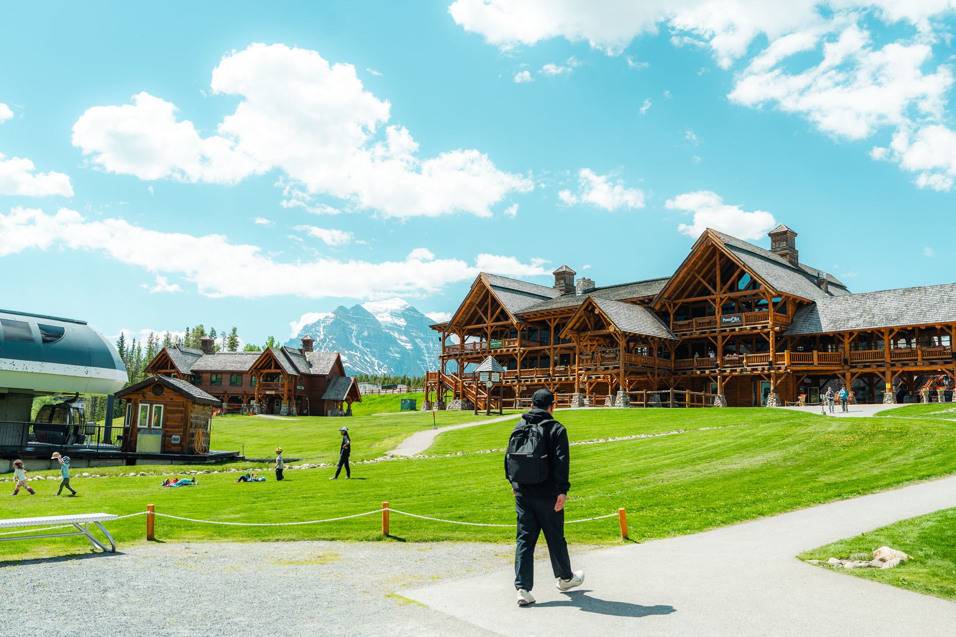 Wooden lodge buildings and ski lift beside grassy field with people walking and playing.