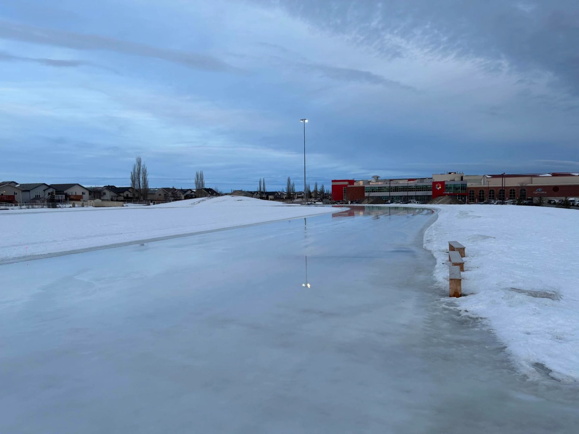 Frozen skating oval with snowbanks, benches, and nearby community buildings.