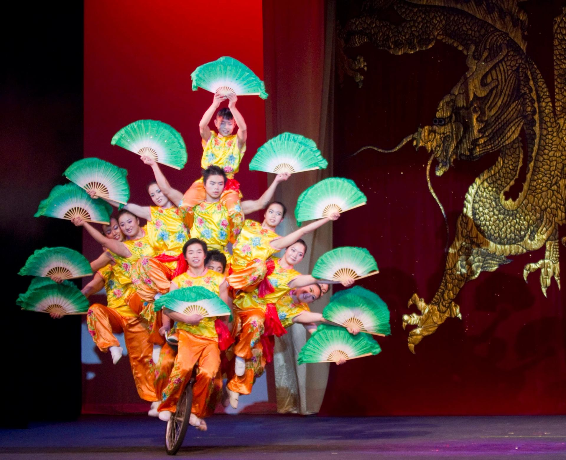 Acrobats in yellow costumes balance on a unicycle, holding green fans before dragon backdrop.
