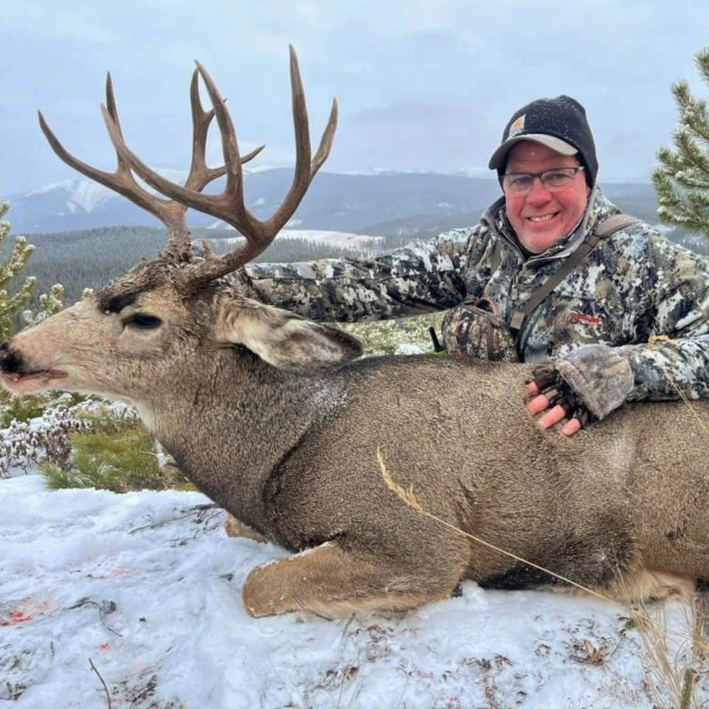 Hunter in camouflage kneels beside large antlered deer on snowy hillside with mountains.