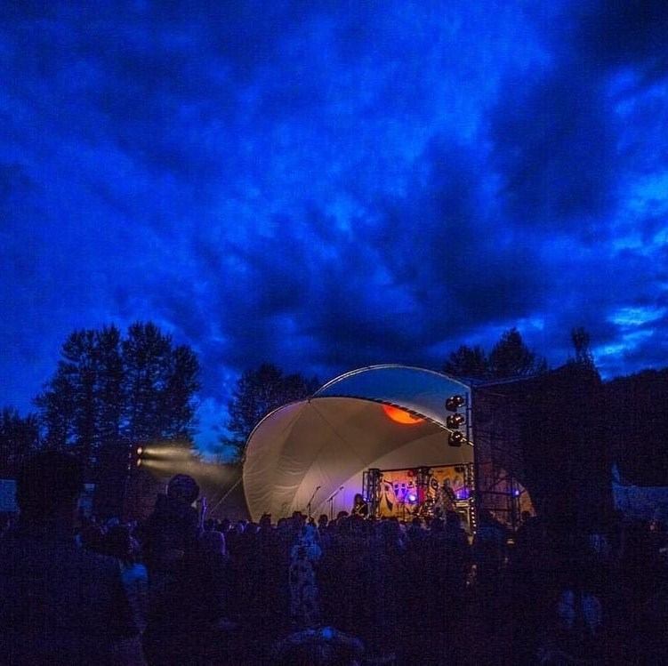 Outdoor stage lit at night under dark blue clouds with a crowd watching a live performance.