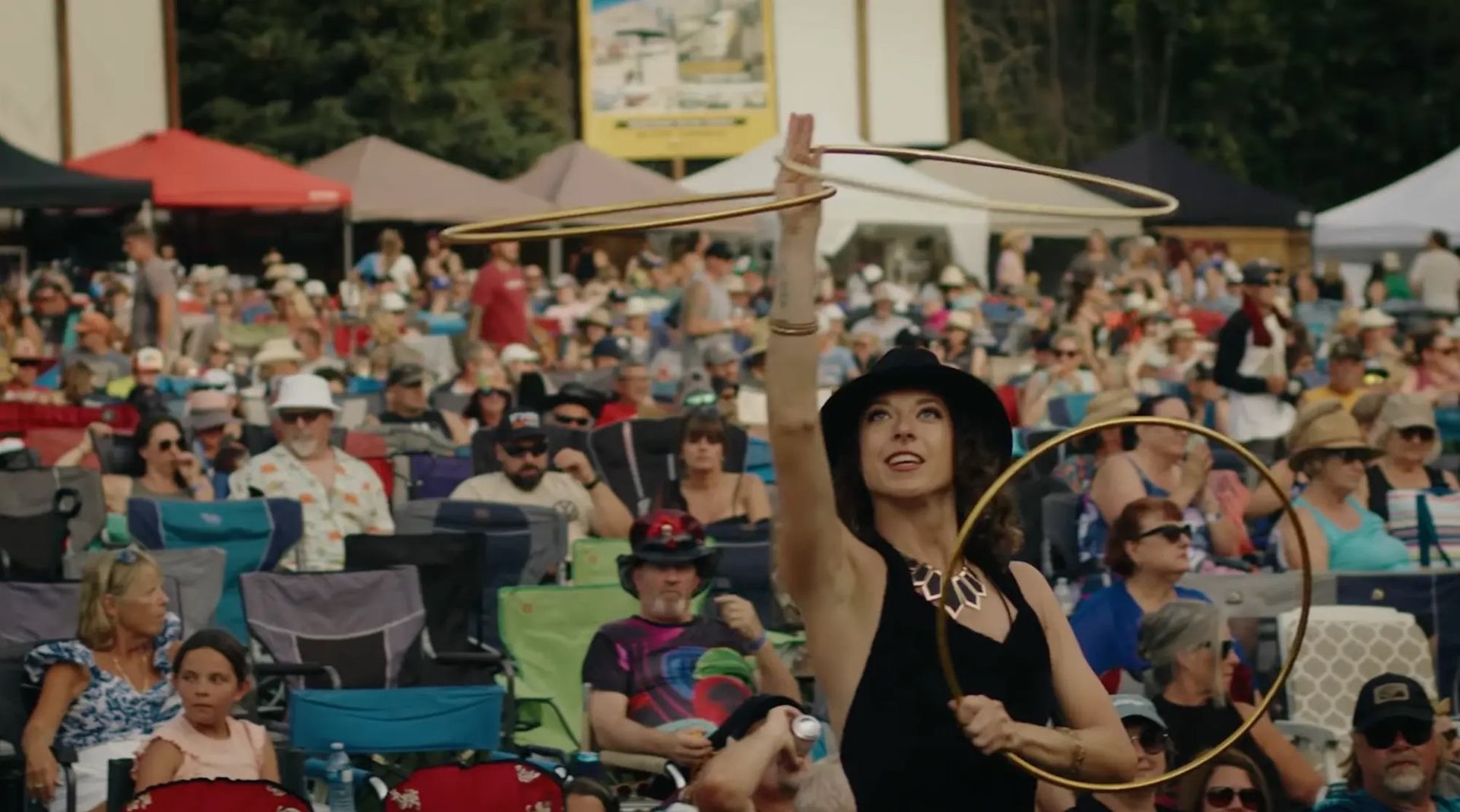 A performer spinning hula hoops in front of a large seated festival crowd outdoors.