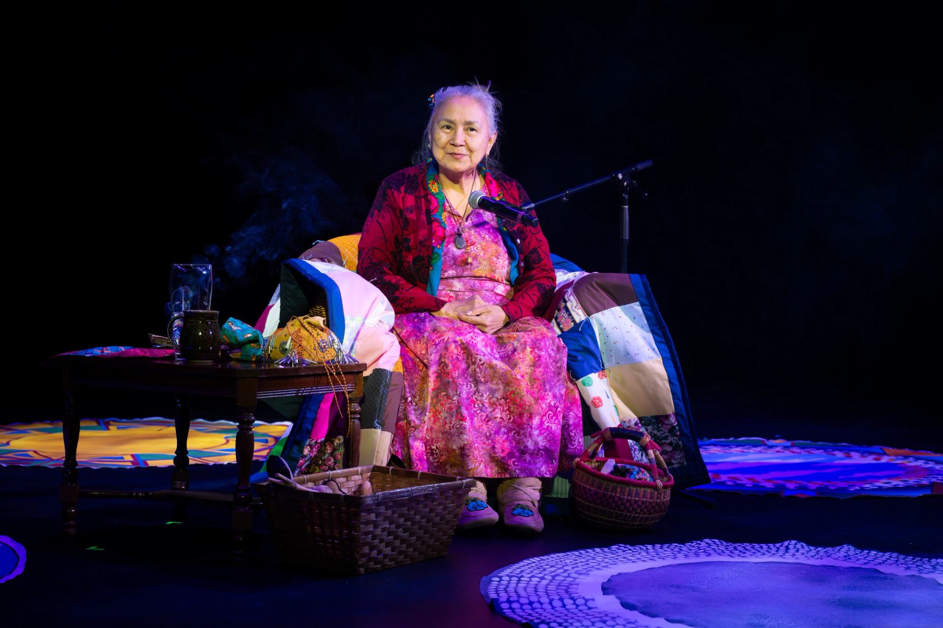 Elder seated in a colorful, traditional setting surrounded by blankets, baskets, and ceremonial items.