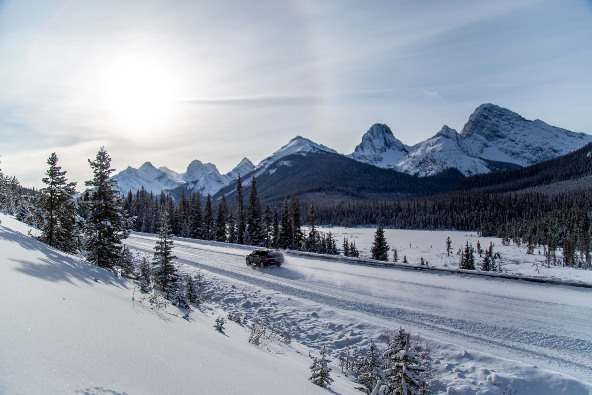 Snow-covered road with a car and mountain peaks under a bright winter sun.