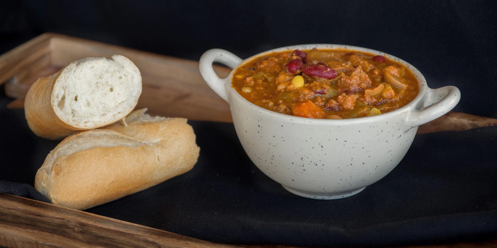 Bowl of chili beside sliced baguette on a wooden tray.