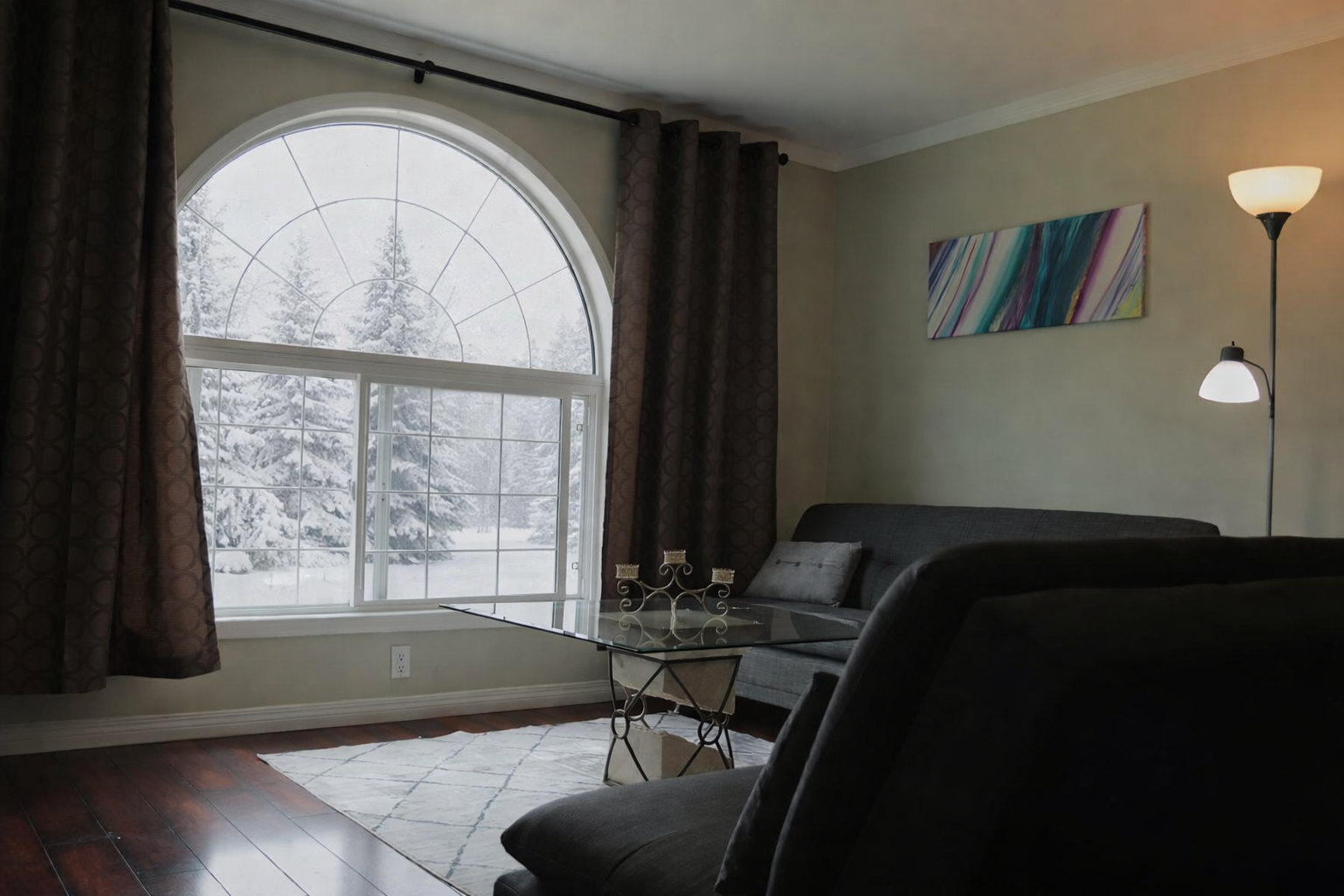 Living room with arched window, snowy trees outside, grey sofa and glass coffee table.