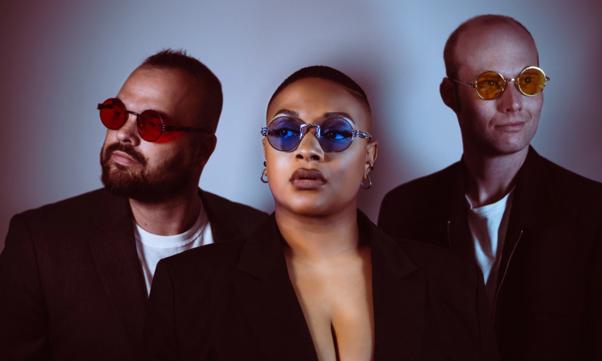 Three people in dark jackets wear coloured sunglasses against a plain gradient background.