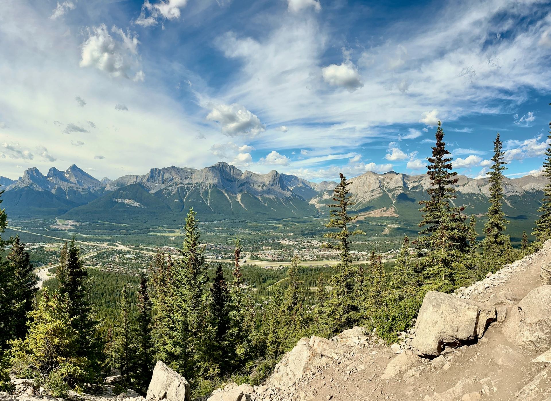 Rocky mountain range above forested valley with town, highway and scattered clouds.