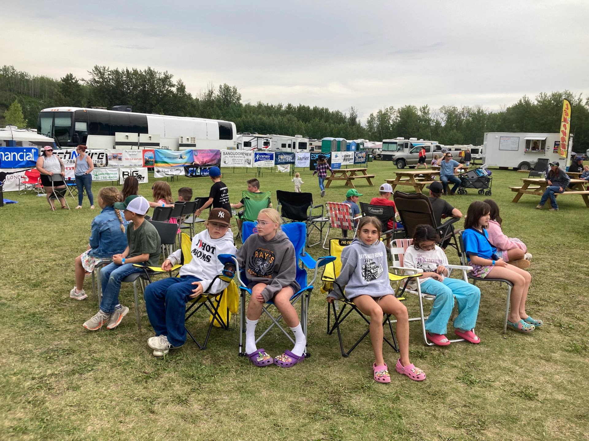Children sit on lawn chairs at an outdoor event with trailers and banners.