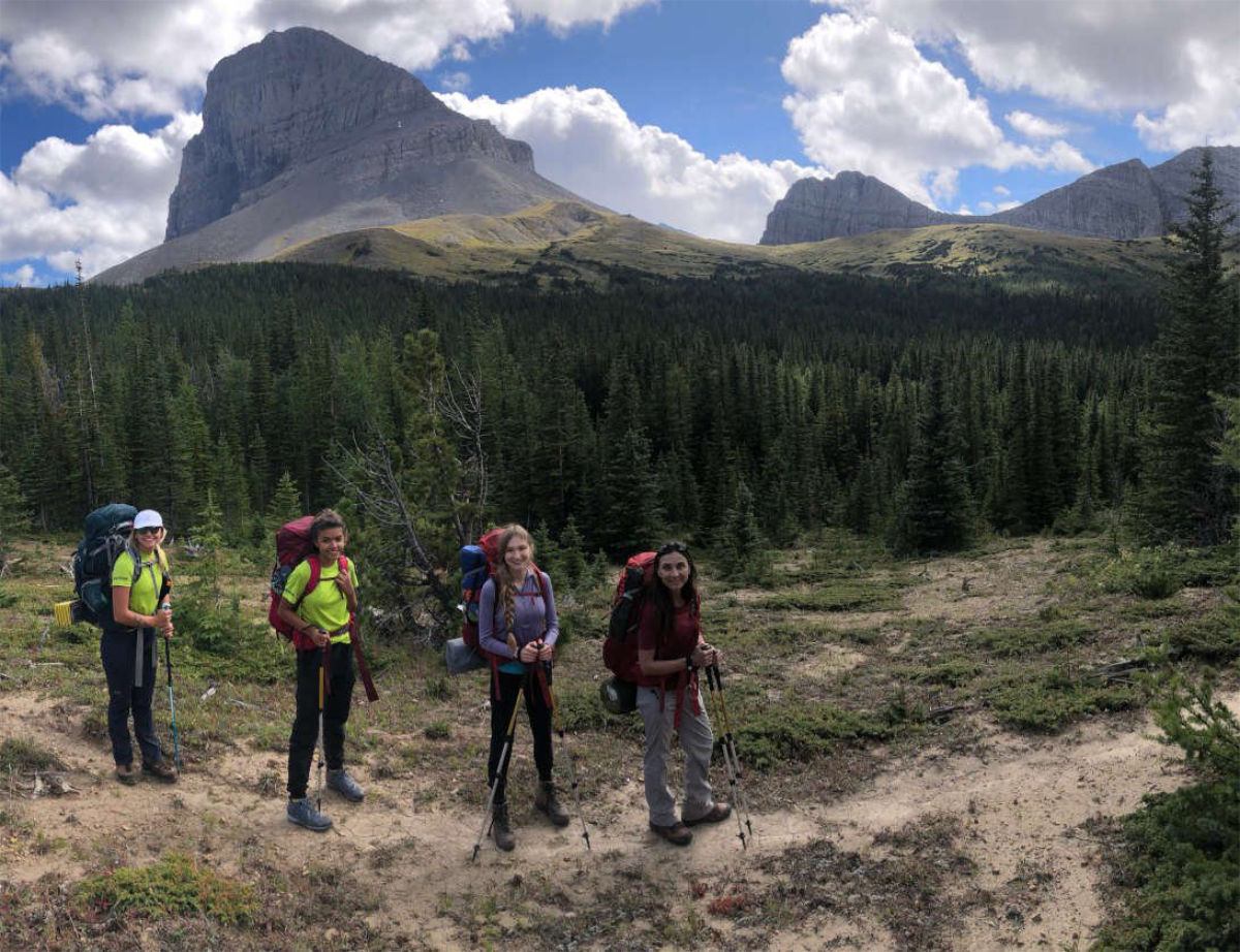 Hikers with backpacks stand on a trail before forested mountains under scattered clouds.