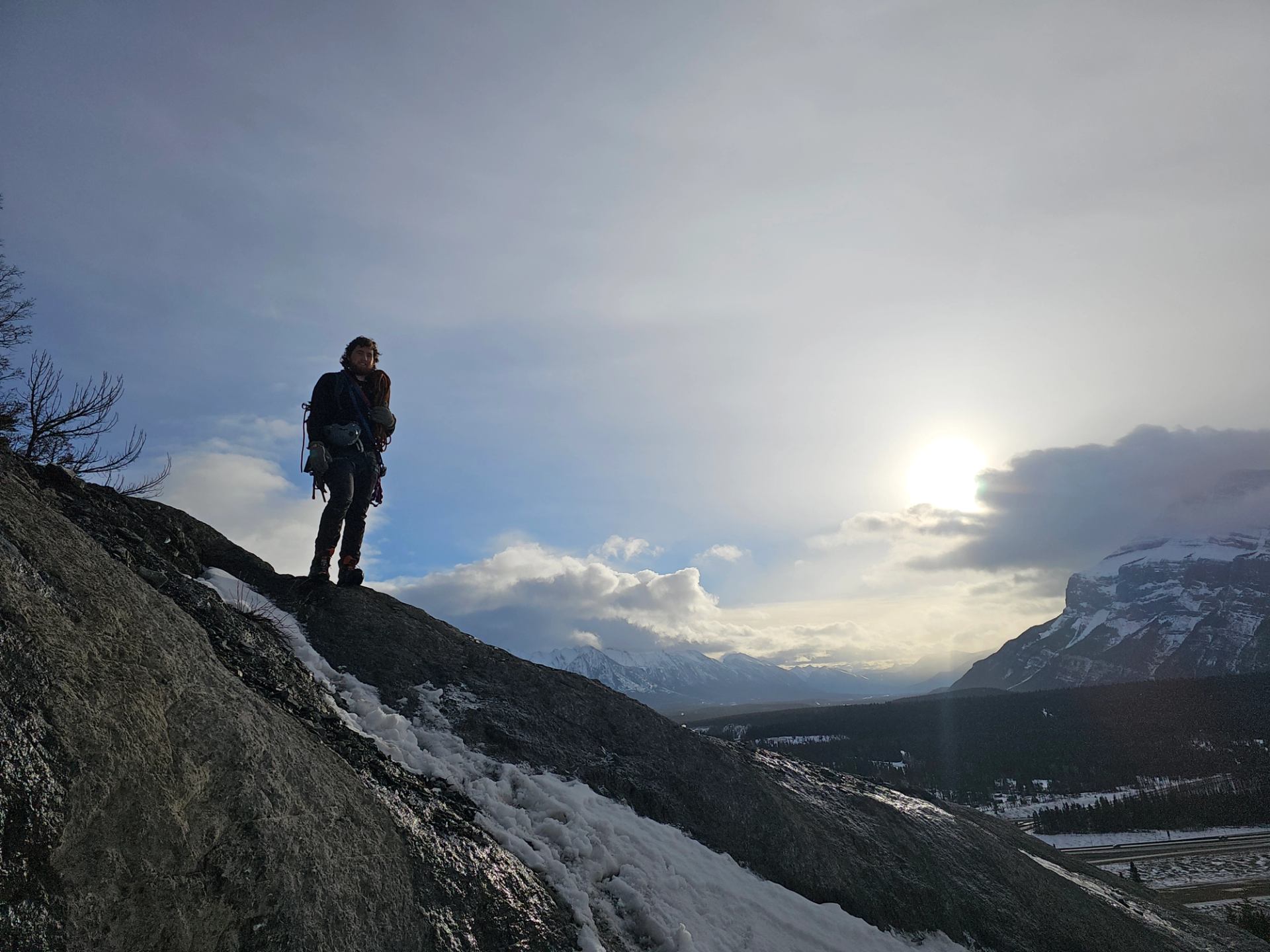 Hiker stands on snowy mountainside trail with backpacks under bright sky.