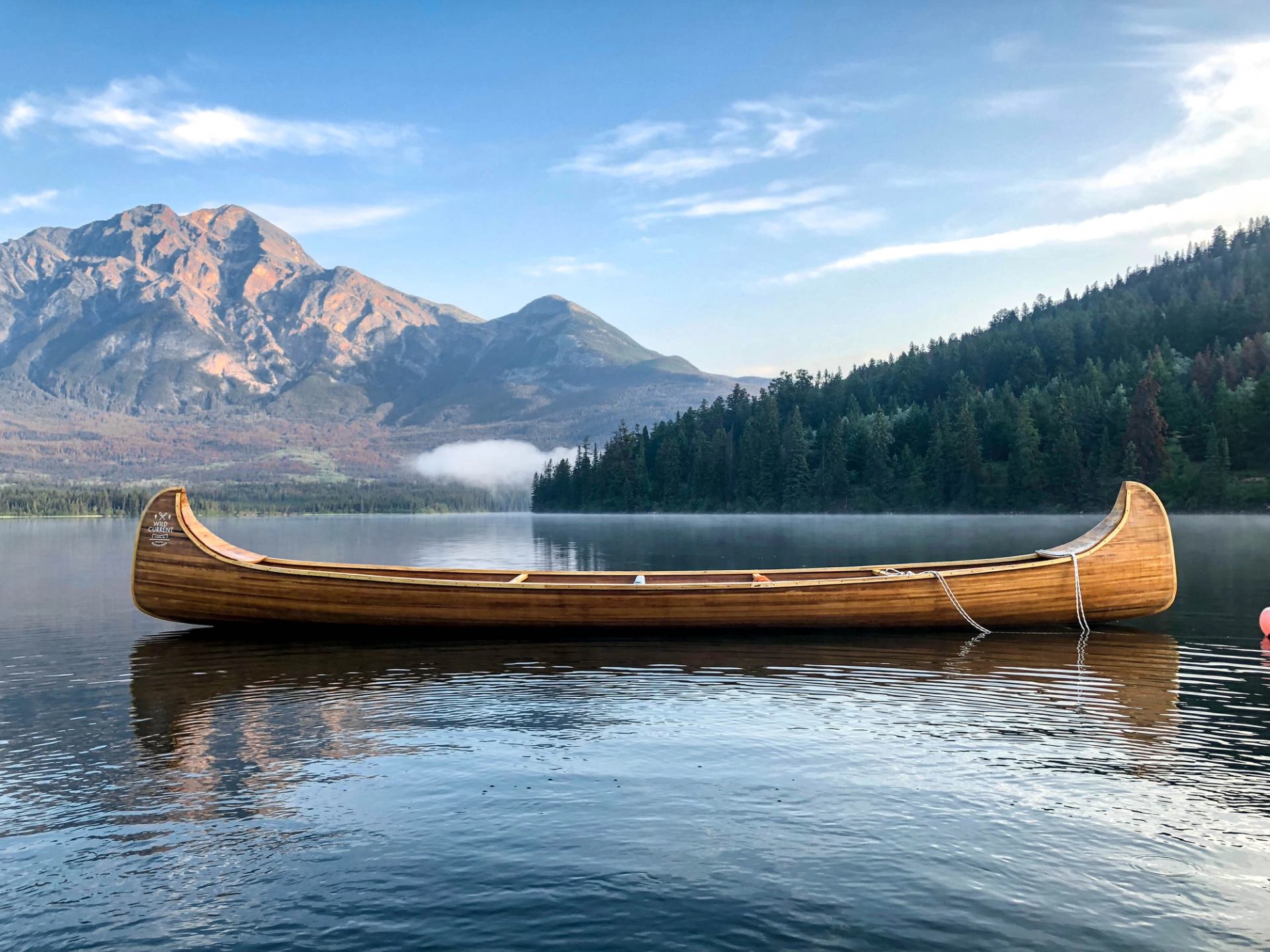 Empty wooden canoe floating on a calm lake with forested shoreline and mountains in the background.