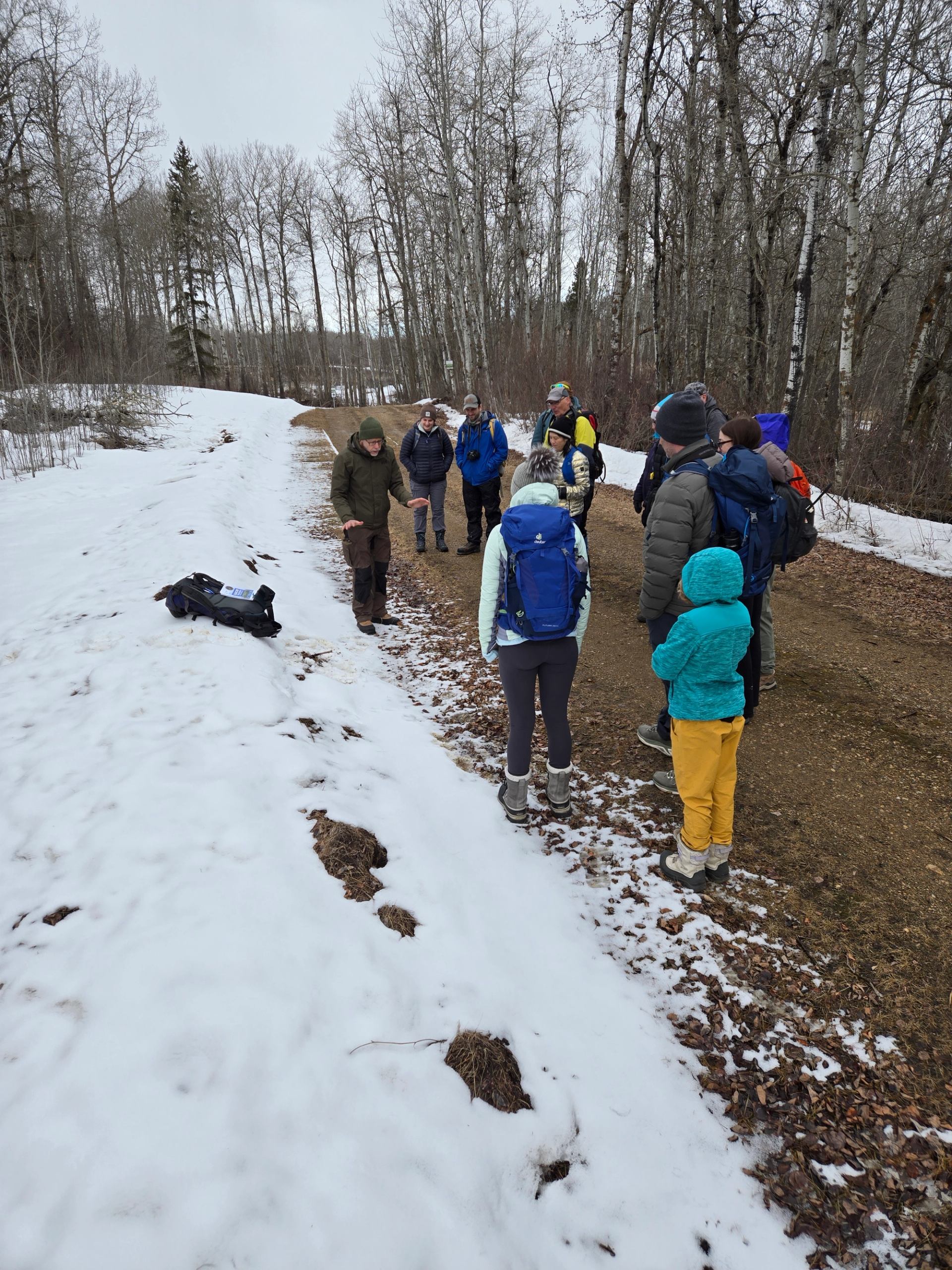Group of hikers standing on snowy forest trail, listening near backpacks on ground.