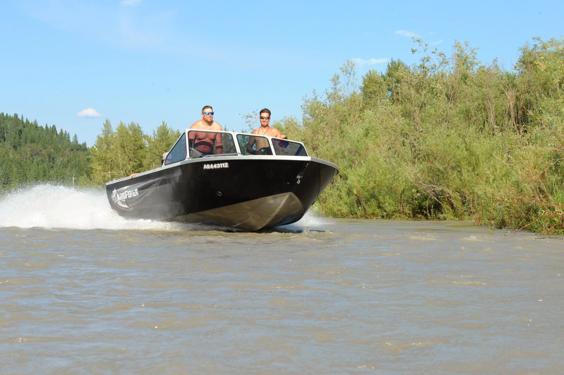 Black motorboat with two riders speeds along a tree-lined river.