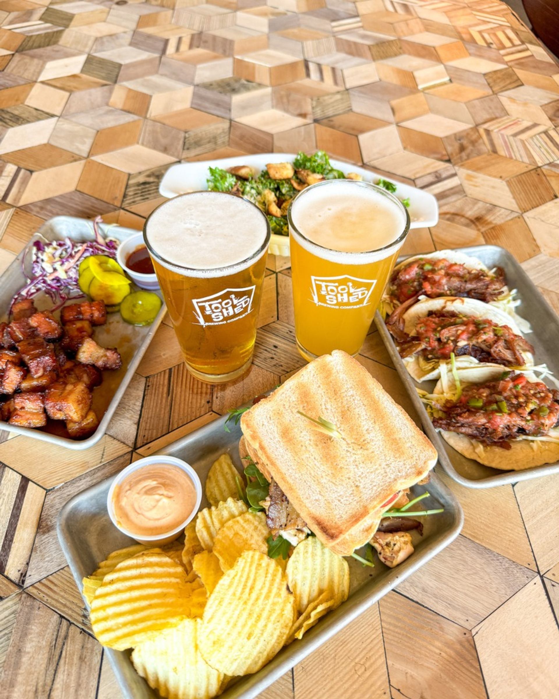 Pub table with two Tool Shed beers, sandwich with chips, tacos and side dishes.