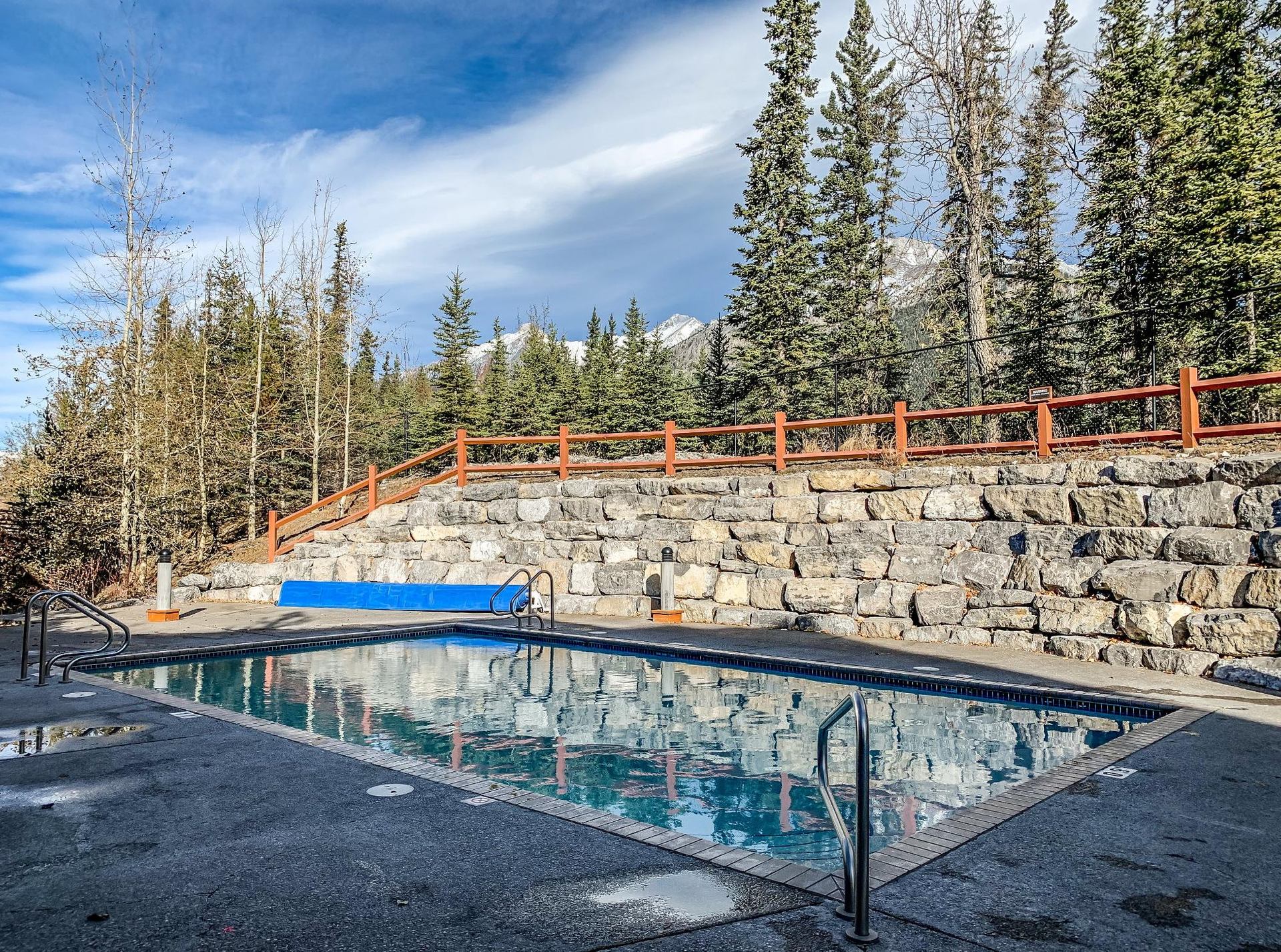 Outdoor swimming pool beside stone retaining wall and evergreen trees under blue sky.